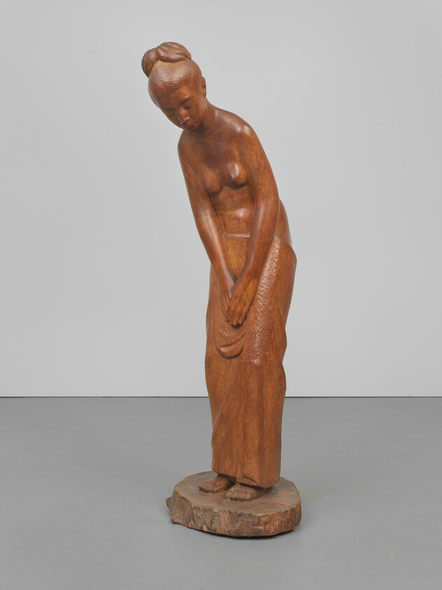 Carved wooden statue of a barefoot woman leaning forward with hands clasped over her skirt.