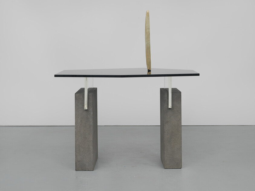 A modern table with a black glass top, supported by two concrete blocks, features a vertical gold accent.