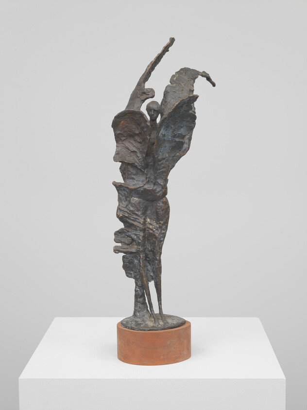 Tall bronze abstract figure with spread wings standing on a round wooden base atop a white pedestal.