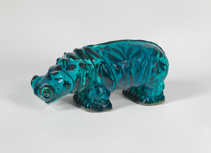 A turquoise glazed ceramic hippopotamus sculpture with dark floral patterns resting on a white surface.