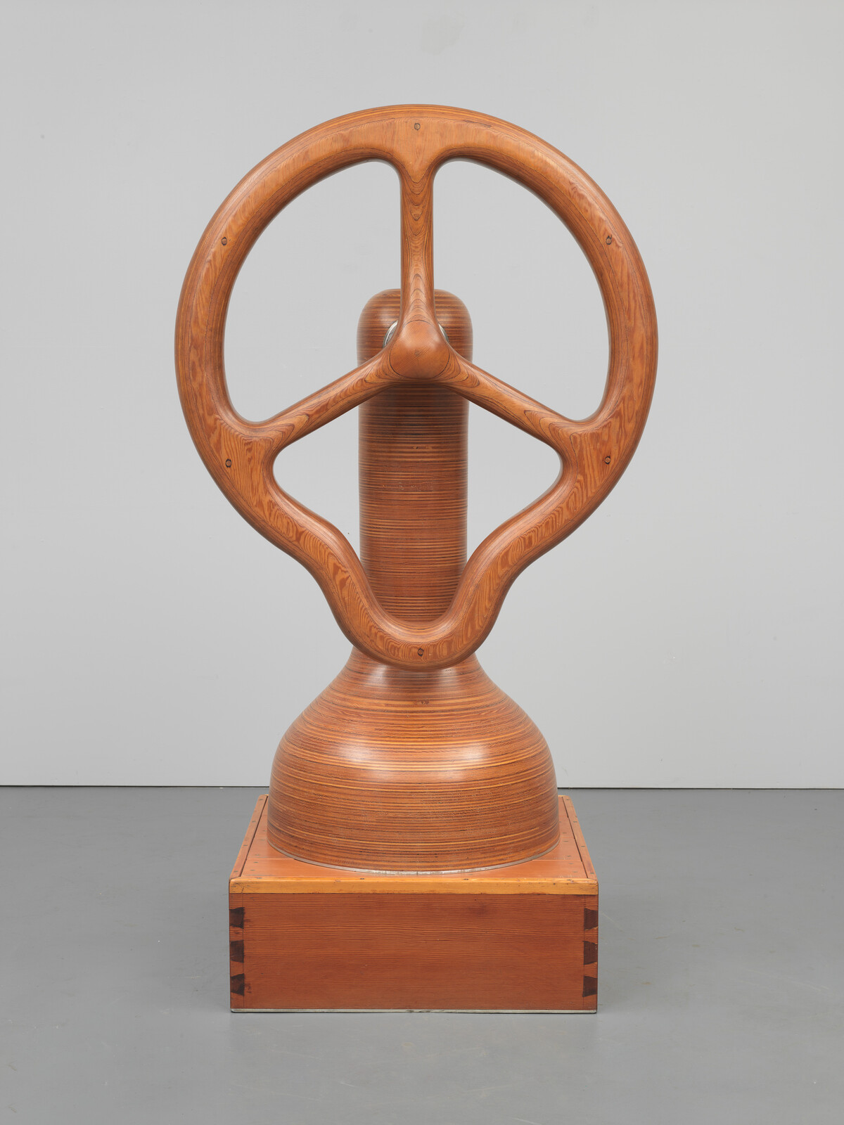 Large polished wooden sculpture resembling a ship wheel mounted on a rectangular wooden base.