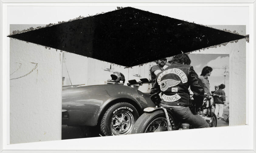 Biker wearing a leather jacket with "Hells Angels France" patch sitting on a motorcycle next to a car.