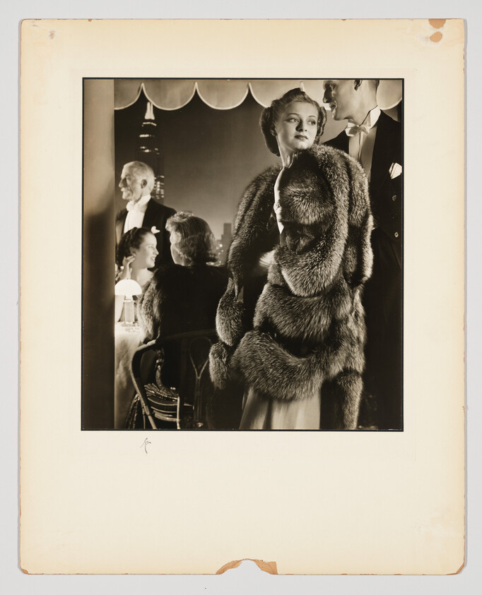 A glamorous woman in a large fur coat stands beside a man in a dimly lit nightclub.