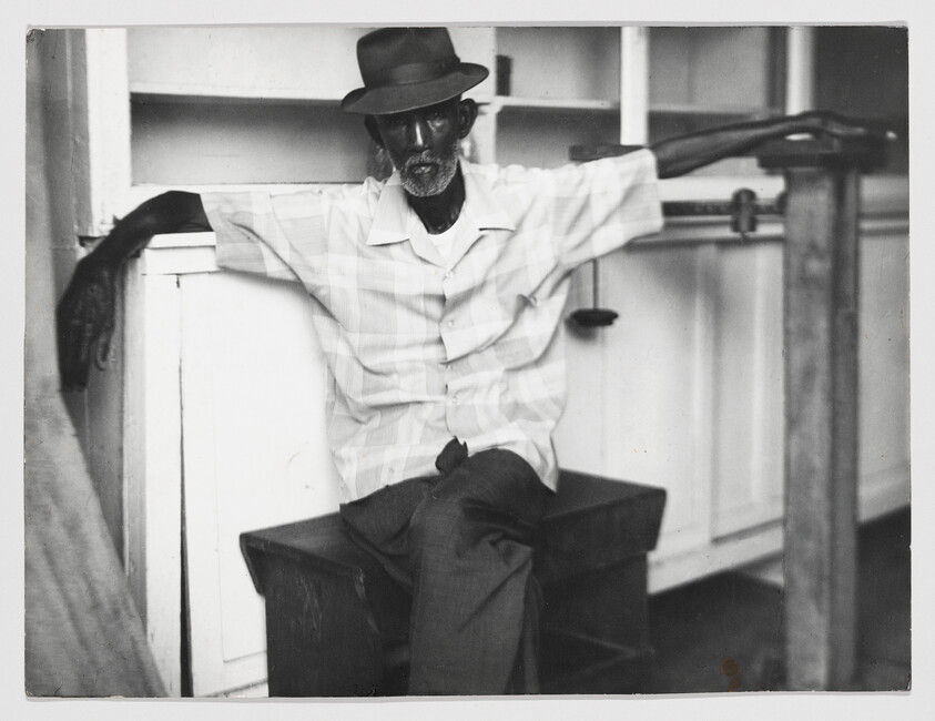 Elderly man wearing a hat sits with arms outstretched across a bench and railing.
