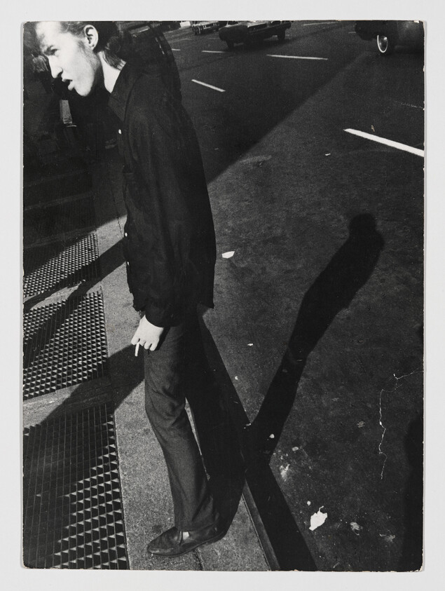 Young person standing on a sidewalk curb holding a cigarette, casting a long shadow across the street.