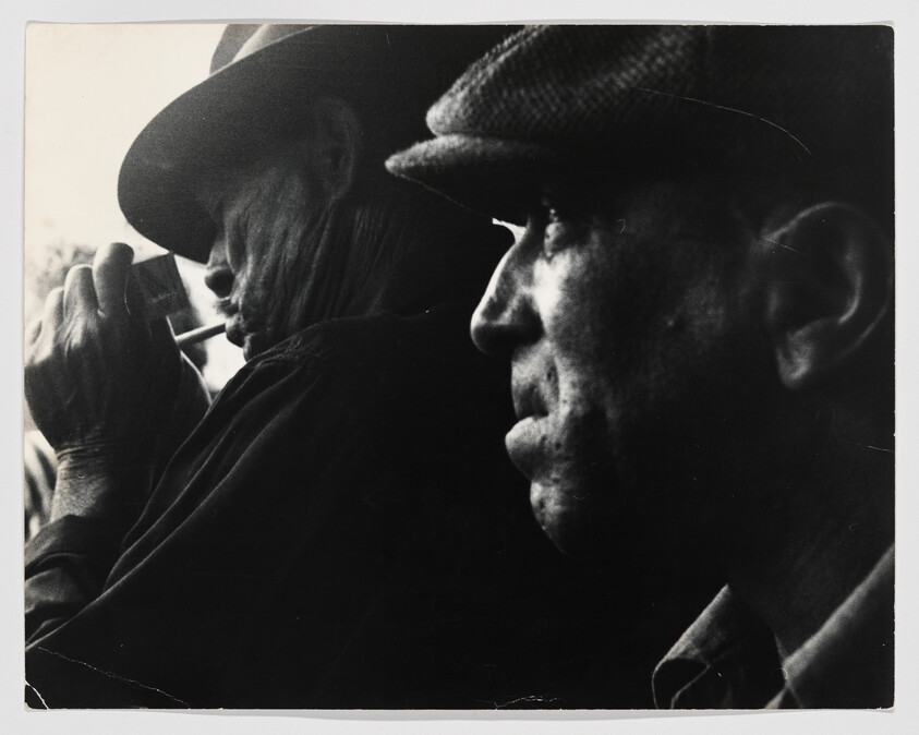 Two men in profile wearing hats, one lighting a cigarette while the other looks ahead.