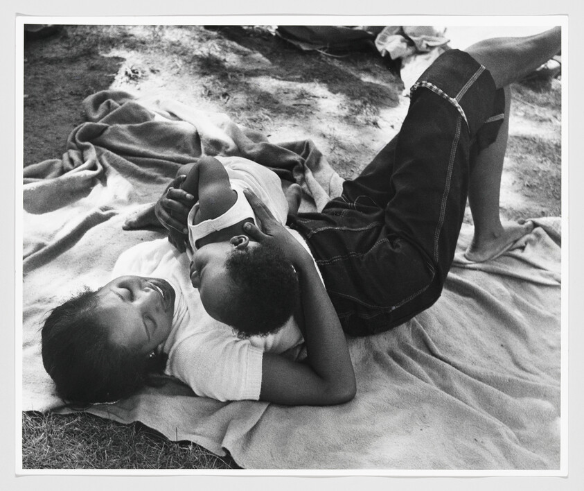 Teen girl lies on a blanket outdoors holding and smiling at a small child cuddled on her chest.