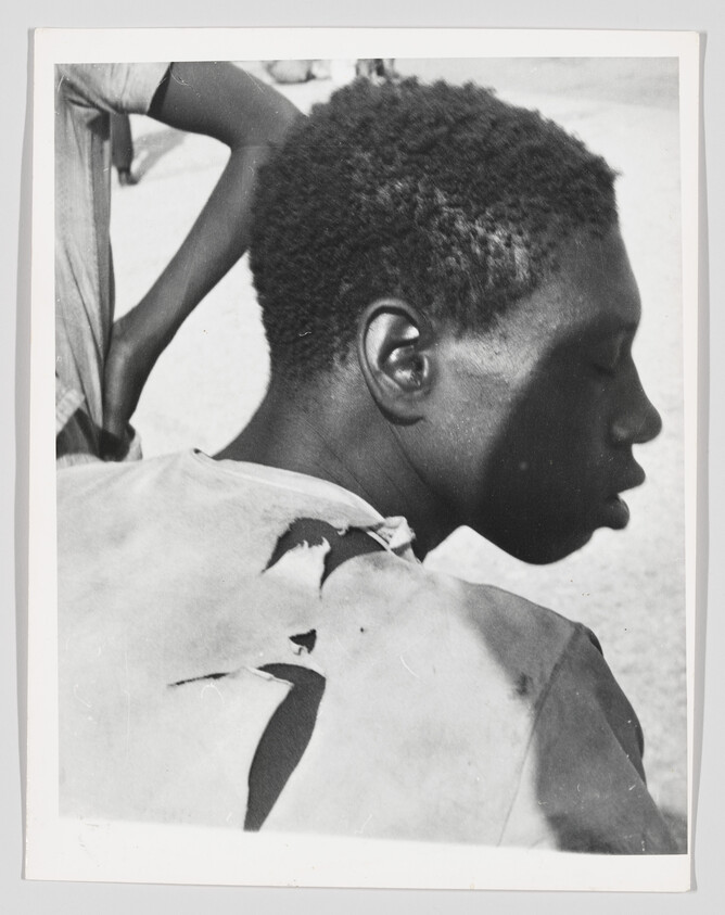 A young person in profile with short curly hair wearing a shirt torn across the back.