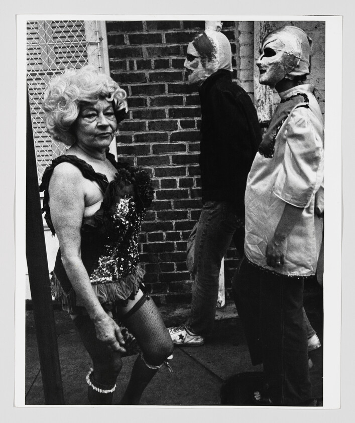 An older woman in a sequined costume walks past two people wearing cloth masks on a sidewalk.