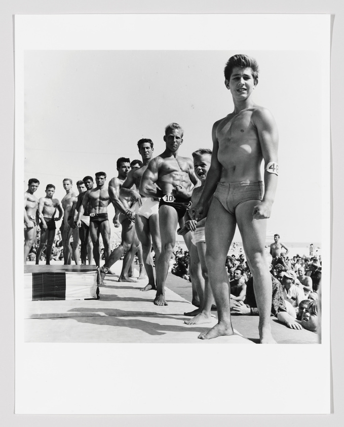 Group of men in swimwear lined up for a competition, with a young man in the foreground smiling at the camera.