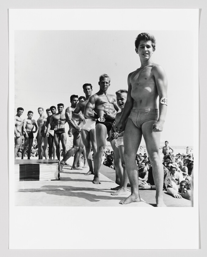 Young men in swim briefs stand in a line on a beach stage during a crowded contest.