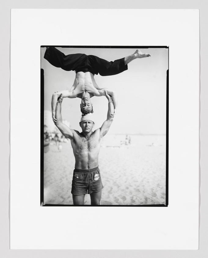 A black and white photograph of a man standing on a sandy beach, holding another man upside down above his head. The man being lifted is in a handstand position, supported by the standing man's outstretched arms. Both men are wearing shorts, and the background shows a clear sky and the hint of a beach environment.