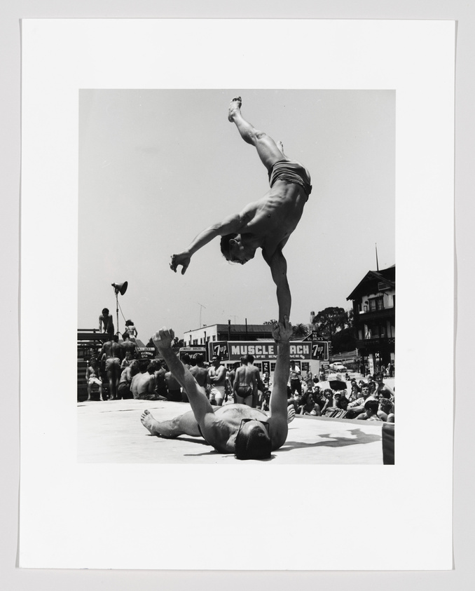 A black and white photograph capturing a moment at a bodybuilding or acrobatic event, with one muscular person balancing upside down on the hands of another who is lying on the ground. The person on top has their arms extended and legs straight up, demonstrating strength and balance. In the background, a crowd of spectators watches the performance, and signs reading "Muscle Beach" are visible, indicating the location of the event.