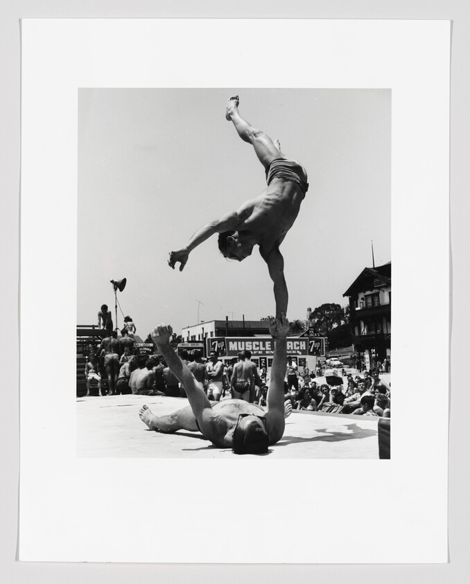 Man lying on his back supports another man performing a one-arm handstand over a crowded outdoor event.