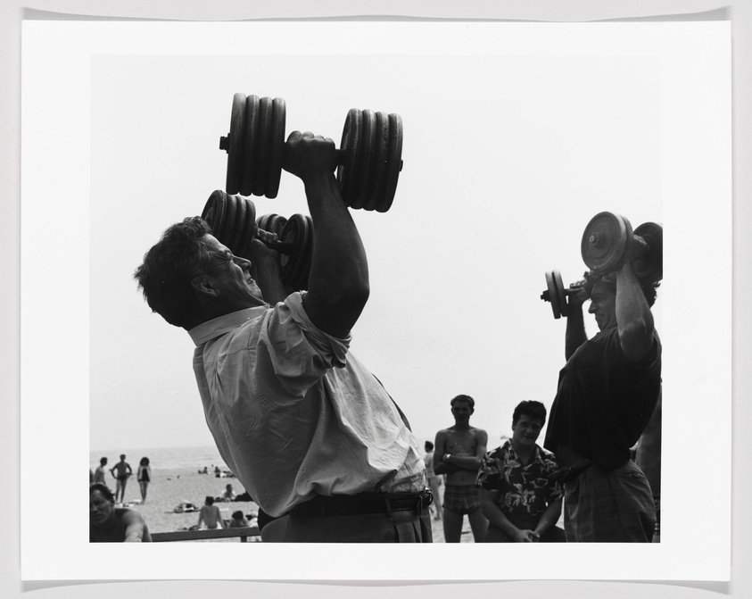 A black and white photograph capturing two men lifting heavy dumbbells overhead at a beach with onlookers in the background.