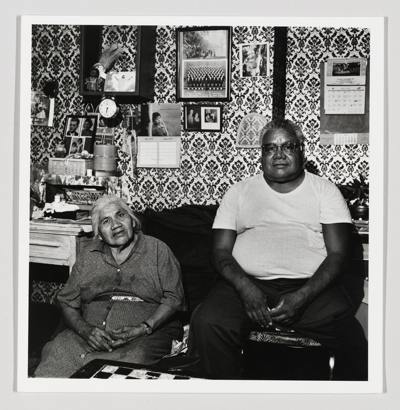 An elderly woman and a middle-aged man sitting in a room with patterned wallpaper and family photos.