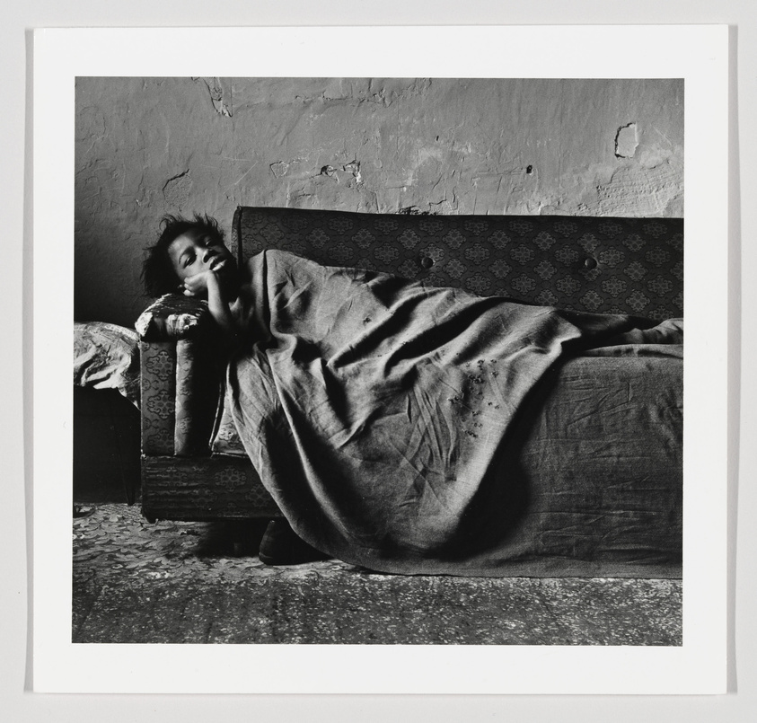 A black and white photo of a person reclining on an old sofa, wrapped in a blanket, in a room with peeling walls.