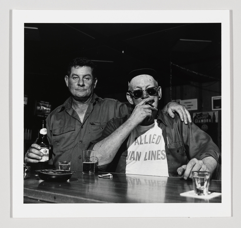 Two men at a bar, one smoking and wearing sunglasses, the other holding a beer bottle.