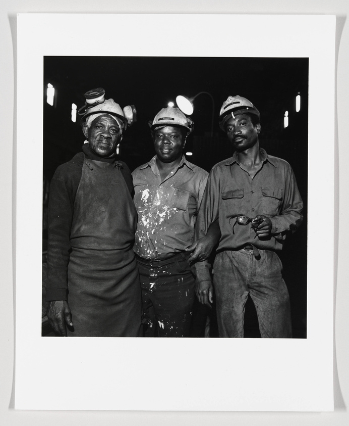 Three coal miners standing close together, facing the camera. They are wearing safety helmets with headlamps and are covered in coal dust. The miner on the left is holding gloves, the one in the middle is smiling slightly, and the one on the right is holding a pair of eyeglasses. The background is dark, highlighting the subjects.