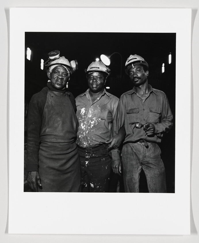 Three coal miners wearing helmets and lamps stand together with arms linked, looking at the camera.