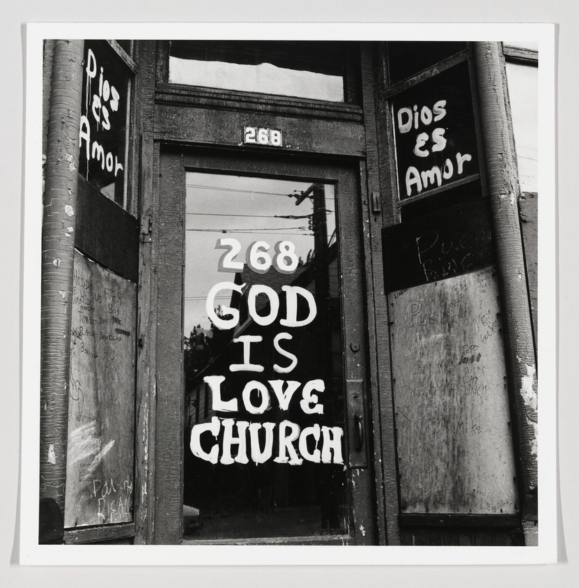 A black and white photograph of a weathered double door entrance to a building with the address "268" above it. The glass doors have "GOD IS LOVE CHURCH" painted in large, bold letters, and graffiti on the surrounding walls includes phrases like "Dios es Amor" and "Puerto Rico."