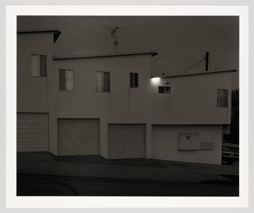 Three closed garage doors under a white apartment building on a sloped street with one light on.