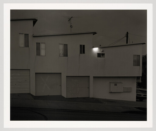 Three closed garage doors under a white apartment building on a sloped street with one light on.