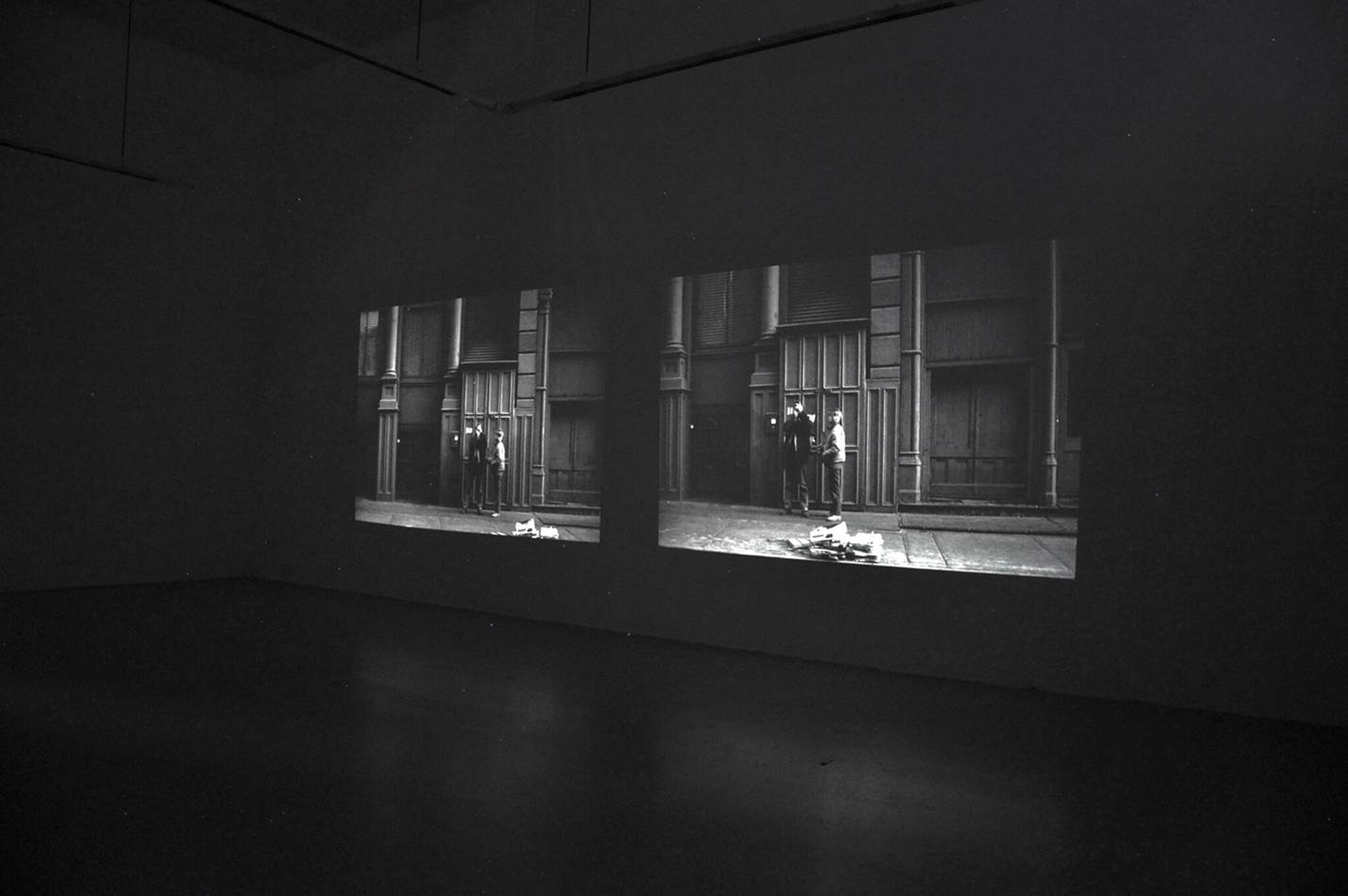 Two black-and-white film projections show people standing by a building entrance in a dark gallery.