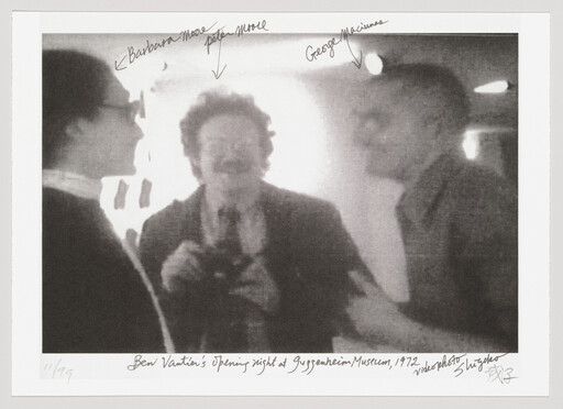 Three people talk at an art opening while a man in the center holds a camera and smiles.