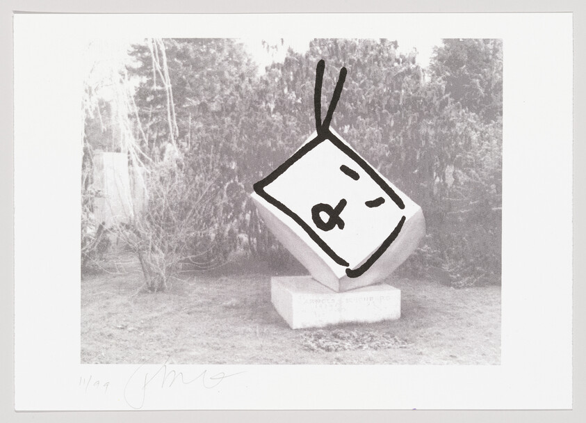 A black and white photograph depicting a whimsical sculpture resembling a large, tilted die with a face drawn on it, including a pair of eyes and a mouth. The sculpture is mounted on a pedestal with an inscription that is partially visible, set against a backdrop of trees and shrubbery. The image also includes a signature in the lower left corner.