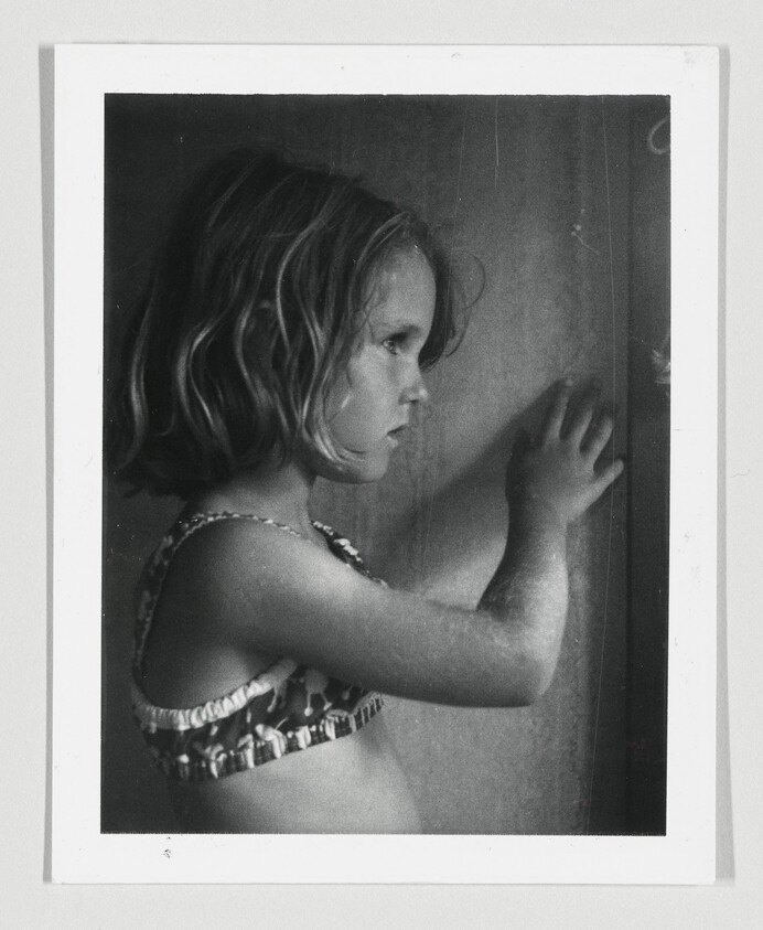 A black and white photograph of a young girl in a patterned swimsuit, standing in profile with her left hand pressed against a dark, textured surface, looking to the side with a contemplative expression.