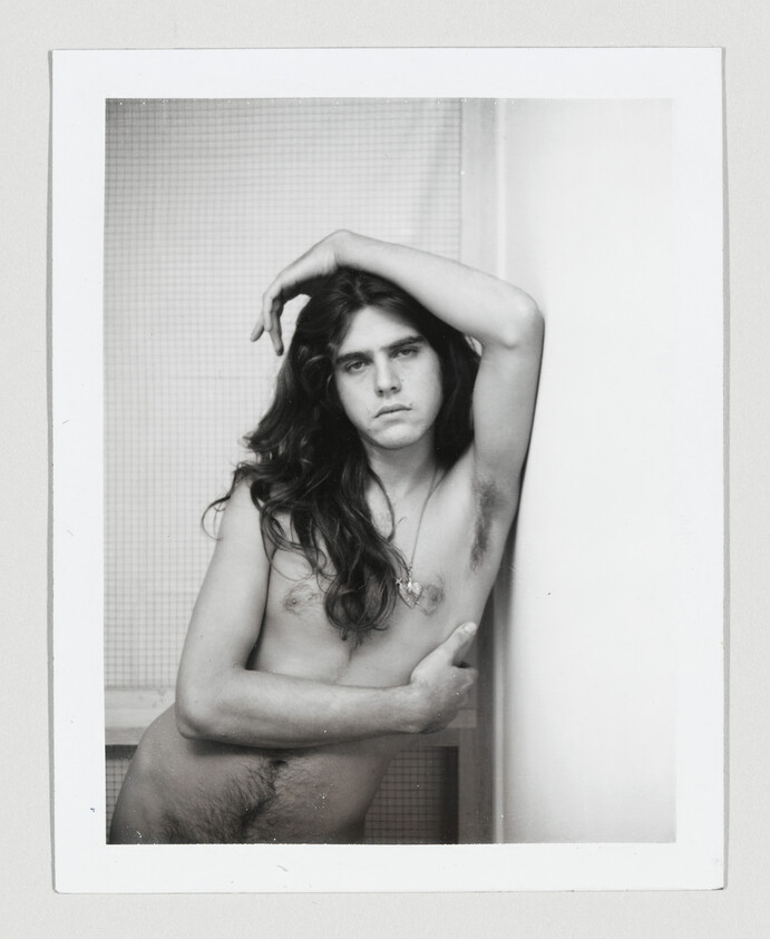 Young person with long hair poses nude against a wall, arm resting above their head.