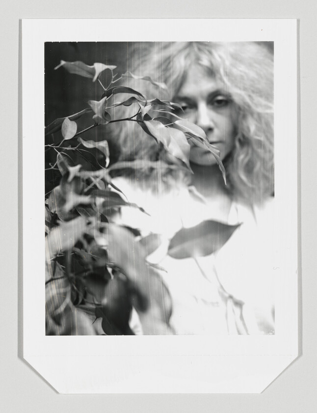 A woman with tousled hair stands behind leafy branches, looking toward the camera.