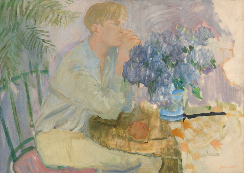 A young man sits at a table, resting his hands by mouth while gazing past a vase of blue flowers.