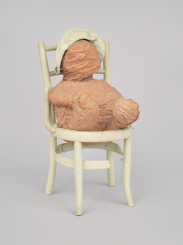 A small white wooden chair holds a wrapped fabric doll wearing a bonnet, viewed from behind.