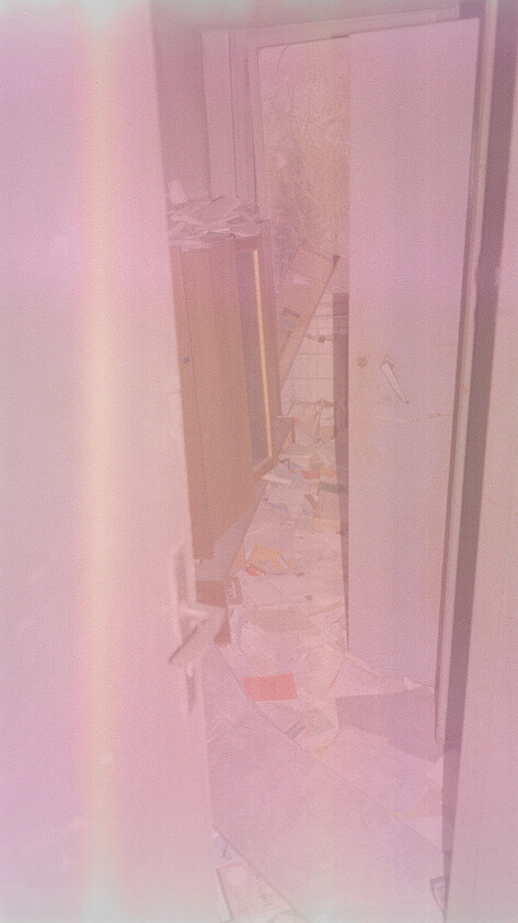 Narrow hallway with open doors and papers strewn across the floor leading to a messy bathroom.