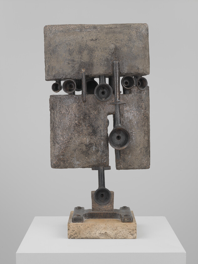 Abstract metal sculpture of stacked rectangular plates and tubular forms mounted on a stone base.