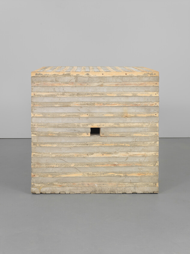 A stacked wooden cube with narrow horizontal slats and a small square opening in front.