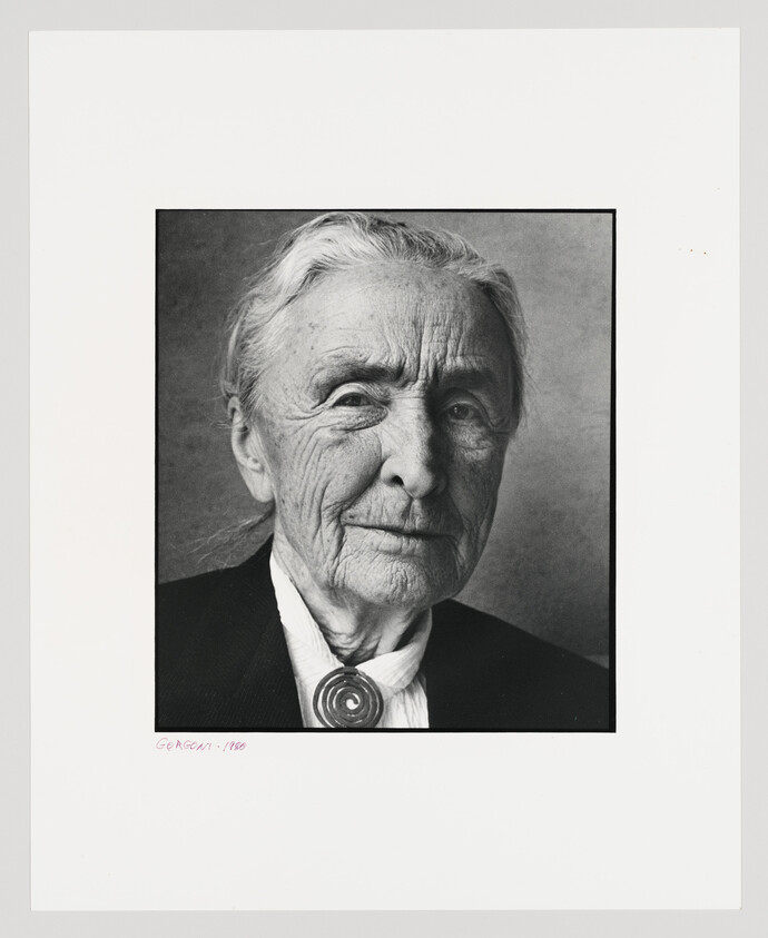 Elderly woman with white hair and deep facial wrinkles wearing a dark jacket and round brooch.