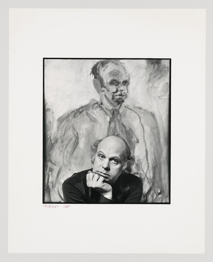 A bald man rests his chin on his hand while sitting in front of a large painted portrait.