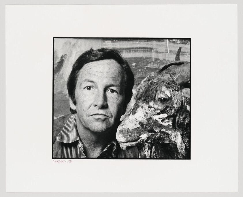 A man poses closely beside a weathered goat sculpture, both facing slightly to the left.