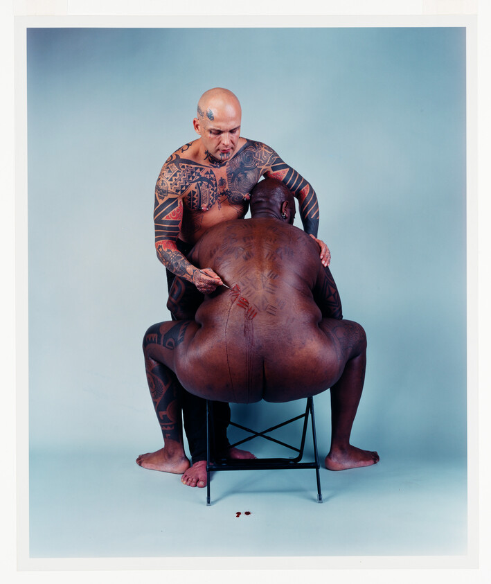 A tattooed man kneels behind another, carving ritual scars into his back while blood drips.