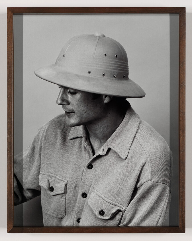 Young man wearing a pith helmet and button-up shirt looks down to his left.