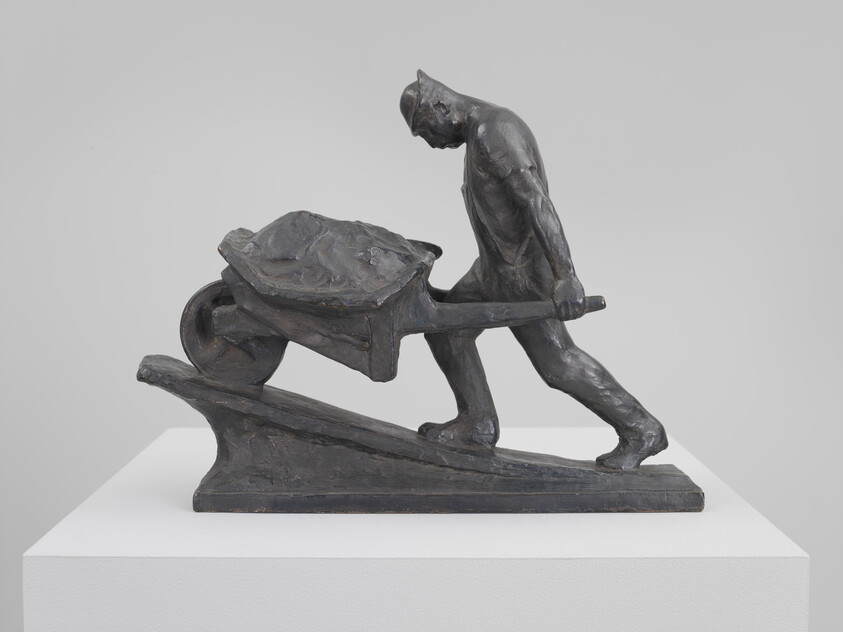 A bronze figure pushes a loaded wheelbarrow up a ramp on a display pedestal.