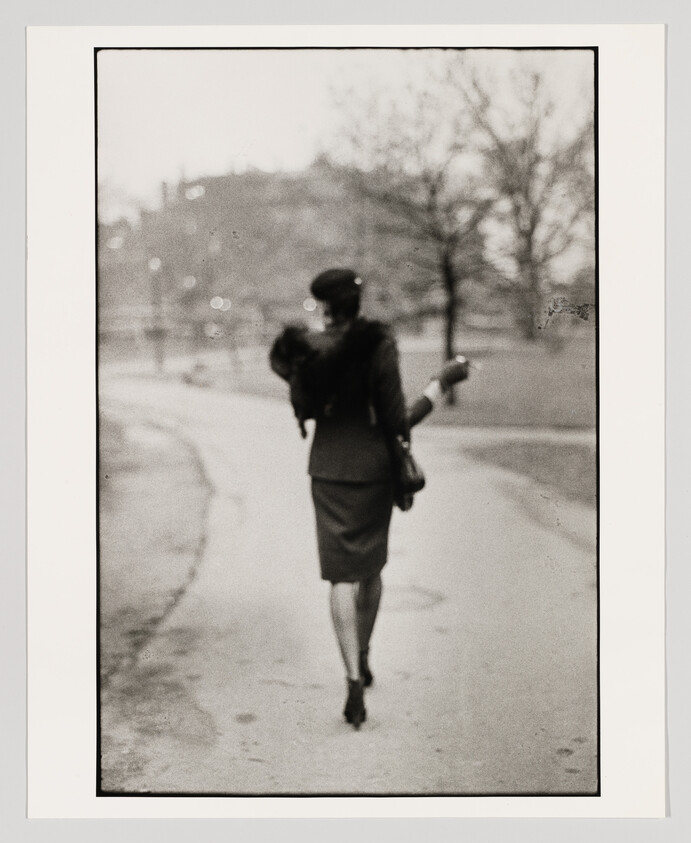 Woman in a vintage suit walks away along a park path, carrying a small dog and handbag.