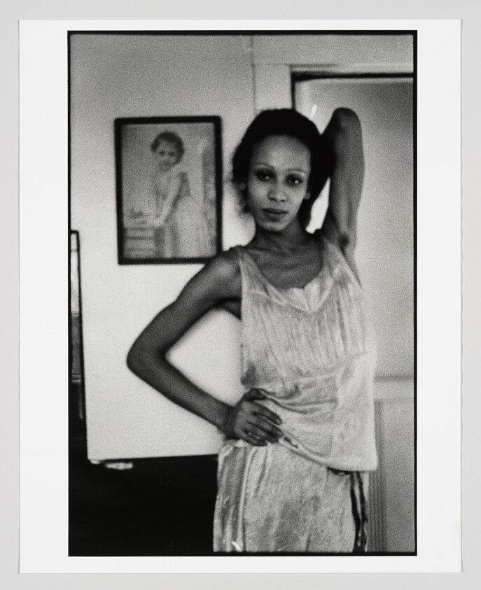 A black and white photograph of a woman standing with one arm raised behind her head, wearing a sleeveless dress. She is looking directly at the camera with a neutral expression. In the background, there is a framed picture of another person on the wall. The image has a grainy texture, suggesting it may be a film photograph or intentionally styled to appear vintage.