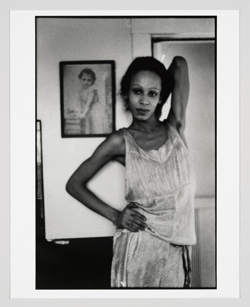 A woman in a sleeveless dress poses with one hand on her hip and the other raised behind her head.