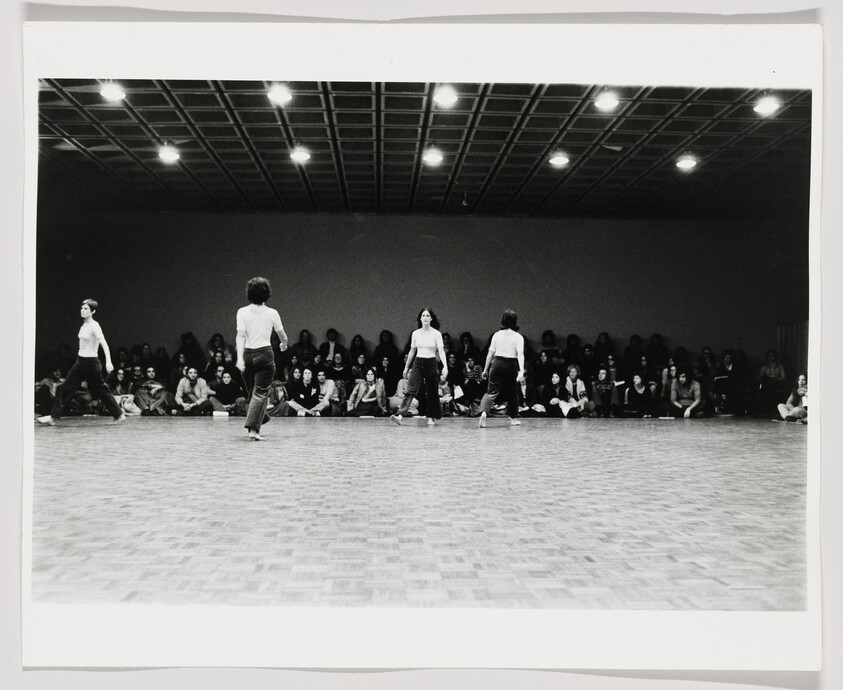 Four dancers walk across a large empty dance floor while a seated audience watches along the back wall.