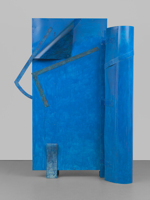 Large blue metal sculpture composed of folded and curved panels standing on a floor.
