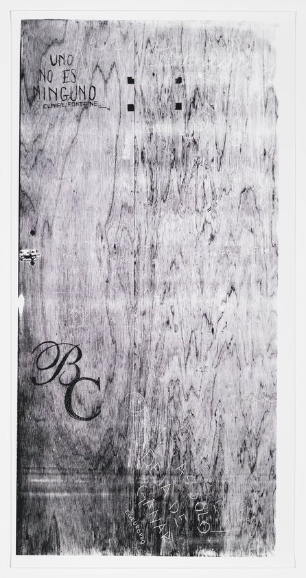 A textured grayscale image of a surface that appears to be a wooden door, with various inscriptions, scribbles, and graffiti in different styles and sizes. Some of the legible text includes "UNO NO ES NINGUNO" and "Claire Fontaine" written in a bold, stylized manner, along with other less discernible writings and marks. The door also features a metal latch on the left side and shows signs of wear and distress throughout.